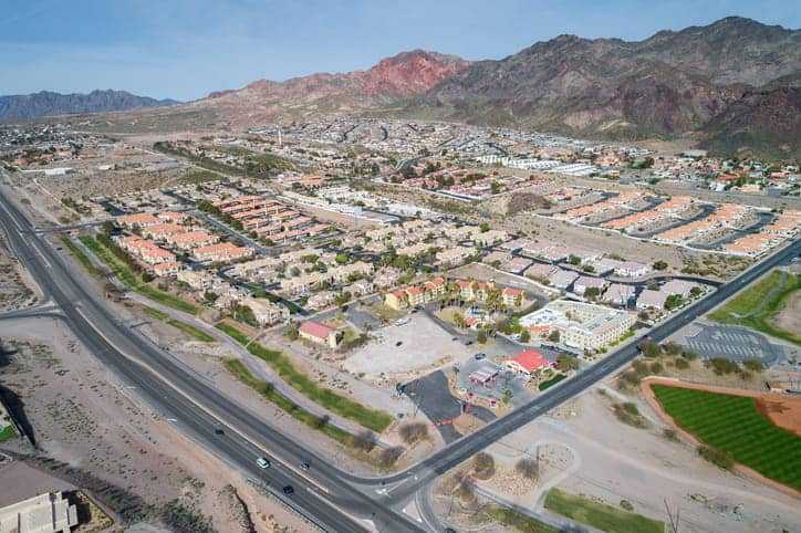Aerial view of a suburban neighborhood in Boulder City NV, with rows of houses and apartment complexes surrounded by roads, parking lots, and desert landscape with mountains in the background under a clear sky—ideal for Boulder City pest control services.