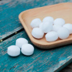 A wooden tray holds several white, round camphor balls—safe alternatives to traditional mothballs—with a few scattered on a weathered, blue wooden surface beside it.