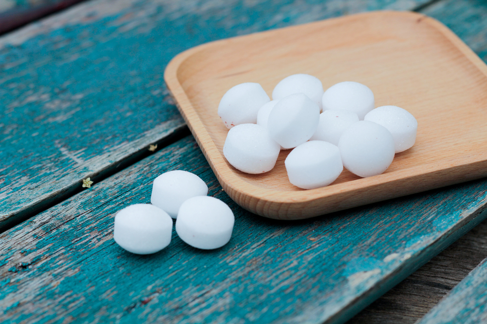 A wooden tray holds several white, round camphor balls—safe alternatives to traditional mothballs—with a few scattered on a weathered, blue wooden surface beside it.