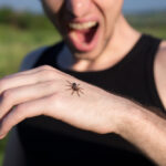 A person wearing a black tank top outdoors looks at a spider crawling on their hand, with their mouth open in apparent surprise or fear—possibly reacting to symptoms of a Yellow Sac Spider bite before considering treatment.
