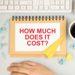 A spiral notebook with "HOW MUCH DOES IT COST?" sits on a desk in Las Vegas, surrounded by a keyboard, coffee cup, plant, hands, binder clips, paper clips, a yellow paper plane, and crumpled paper balls—perfect for pondering pest control cost.