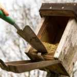 A person uses a small garden trowel to clean out an old wooden birdhouse with a hinged roof. The birdhouse appears weathered and mounted on a metal pole outdoors.