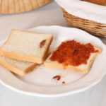 Two slices of white bread on a white plate, one with red spread and the other plain, both topped with a light brown cockroach. A basket of bread rolls is visible in the background—perfect for identification or learning control tips.