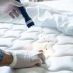 A person wearing gloves inspects a mattress with a flashlight, illuminating several small bugs on the surface. The bed sheet is partially pulled back, exposing the mattress.