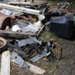 A pile of assorted scrap metal, rusty pipes, a bicycle wheel, old electronics, and various discarded objects scattered on a paved surface with some weeds growing nearby—perfect conditions for spider attraction.