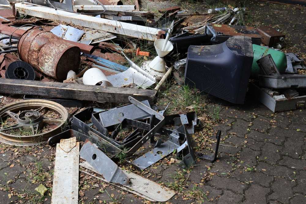 A pile of assorted scrap metal, rusty pipes, a bicycle wheel, old electronics, and various discarded objects scattered on a paved surface with some weeds growing nearby—perfect conditions for spider attraction.